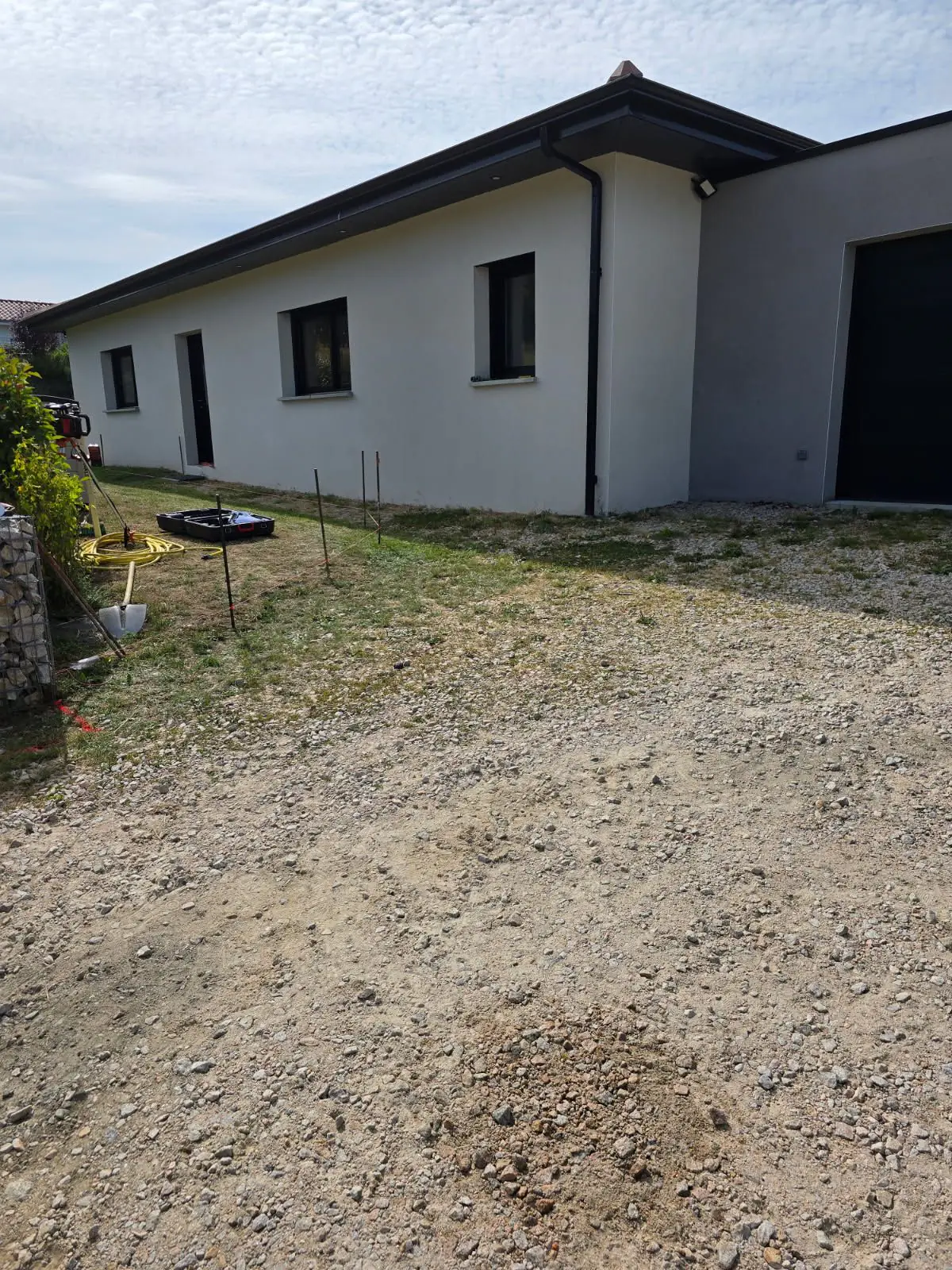 Avant travaux : abords en gravier d'une maison plain-pied blanche, piquets d'implantation en place