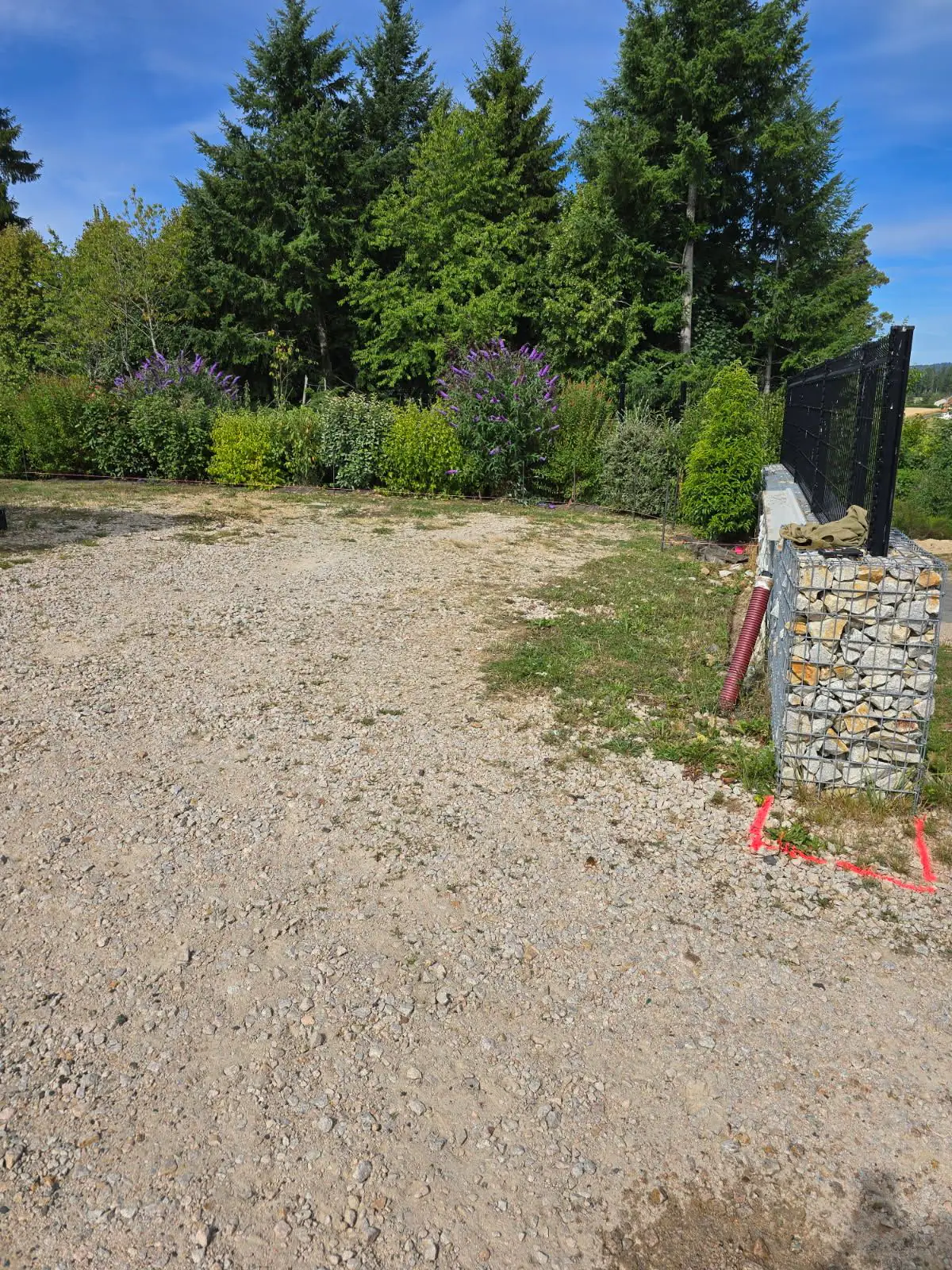 Avant travaux : accès en gravier le long d'un muret en gabions, haie fleurie en fond