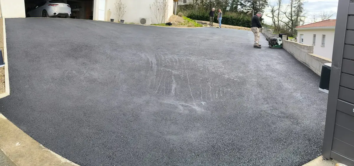 Cour en enrobé neuf devant une maison contemporaine, un ouvrier compacte les bords à la plaque vibrante