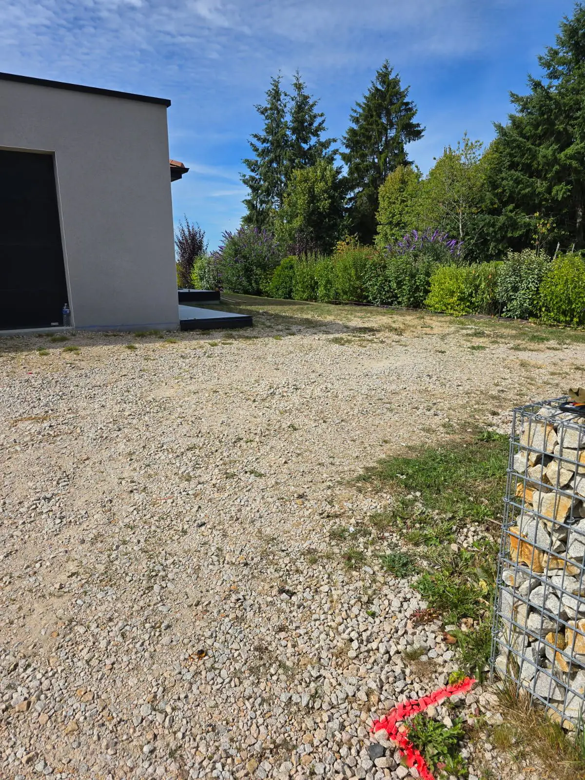 Avant travaux : cour en gravier devant un garage contemporain, haie fleurie et gabion