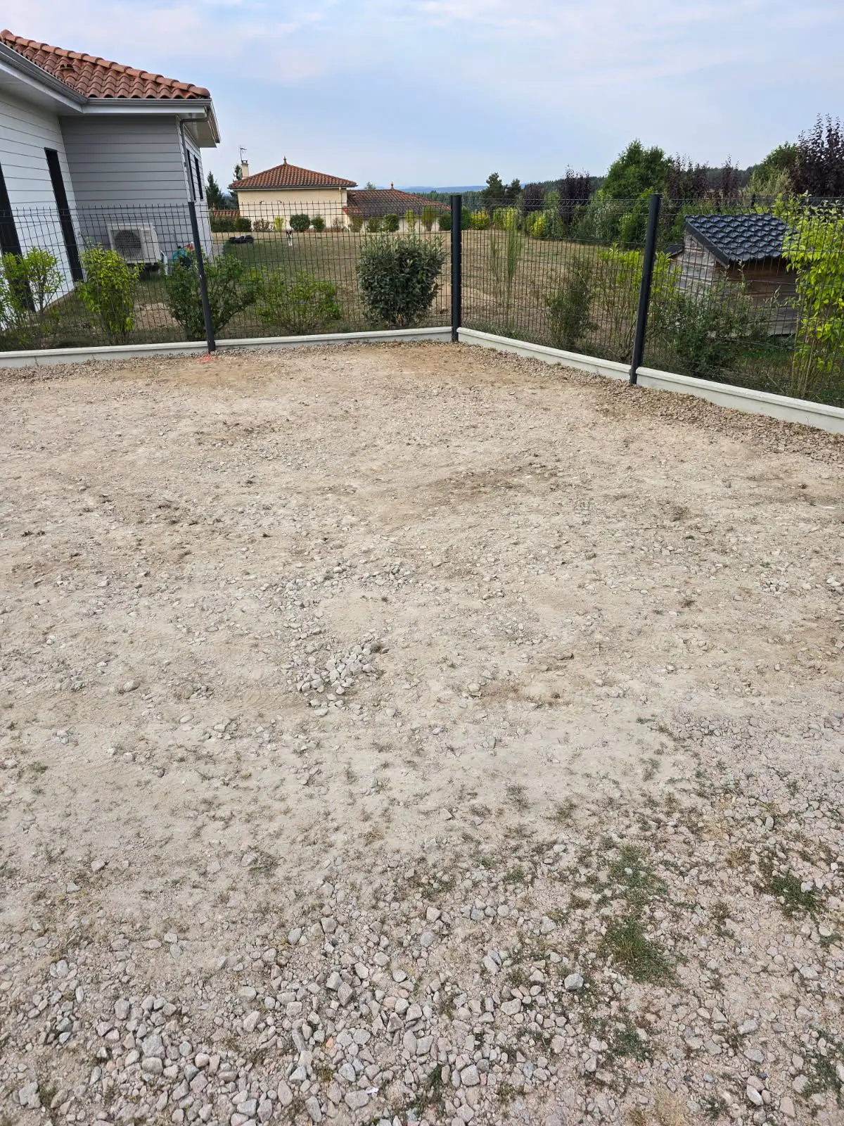 Avant travaux : plateforme en gravier clôturée à côté d'une maison à bardage blanc