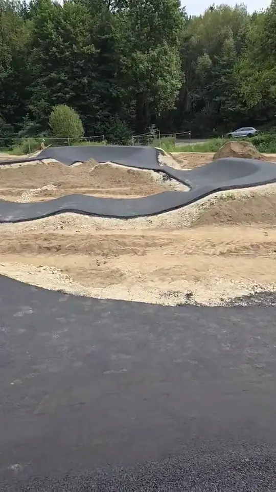 Pumptrack en enrobé noir dans un vallon boisé, bosses et virages relevés, engins de chantier au fond